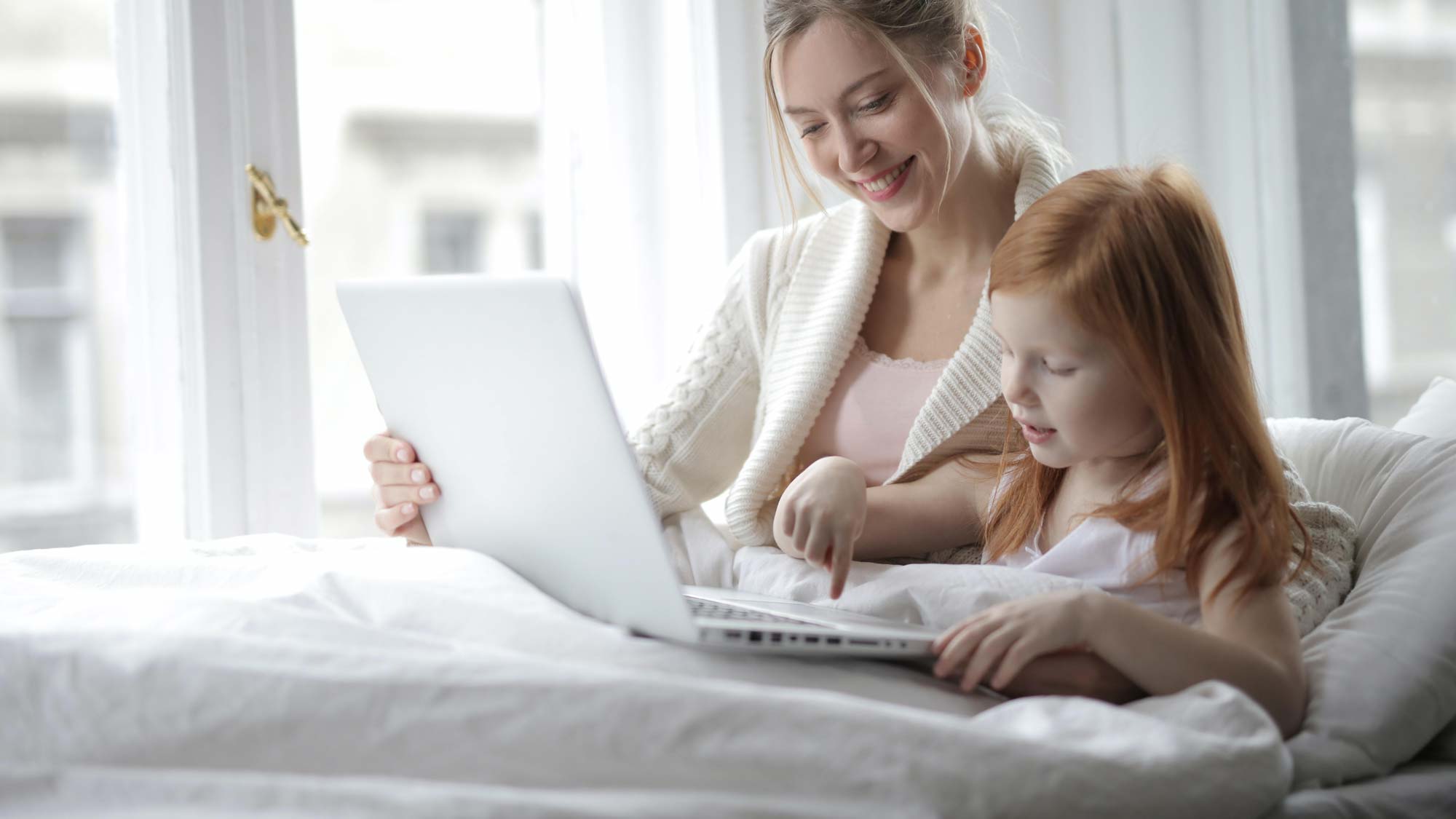 Adult and child sitting on a bed using a laptop together in a bright room.